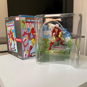 May include: Iron Man action figure in a clear plastic display case. The figure is red and gold, posed on a green base. A box with comic book art featuring Iron Man is visible to the left. The box has the Marvel Comics logo and the words "The Invincible Iron Man."