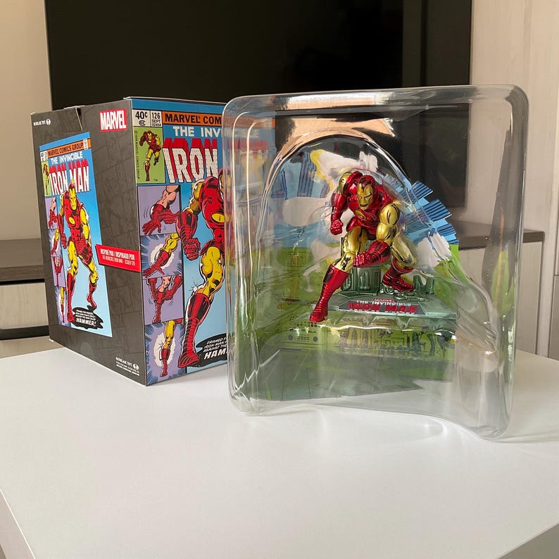 May include: Iron Man action figure in a clear plastic display case. The figure is red and gold, posed on a green base. A box with comic book art featuring Iron Man is visible to the left. The box has the Marvel Comics logo and the words "The Invincible Iron Man."