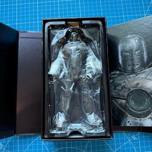 Może przedstawiać: Srebrna figurka Iron Mana w czarnym pudełku. Figurka jest zamknięta w przezroczystym plastiku. Pudełko jest otwarte, ukazując figurkę i boczny panel ze szczeg&oacute;łową ilustracją hełmu Iron Mana.