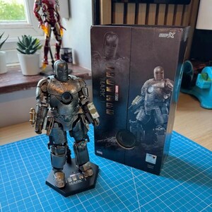 Może przedstawiać: Szczeg&oacute;łowa figurka Iron Man Mark 1 w kolorach szarości i brązu, stojąca na czarnej podstawie z napisem "MARK 1". W tle widoczne pudełko z napisem "IRON MAN" i "MARVEL STUDIOS".