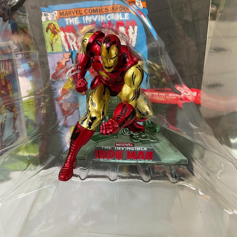 May include: A red and gold Iron Man action figure in a dynamic pose. The figure is on a base with the text "Marvel The Invincible Iron Man". The background features a comic book cover.