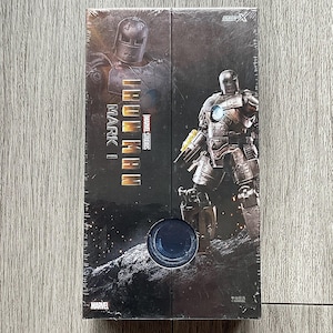 Może przedstawiać: Zapakowane pudełko z Iron Man Mark I z Marvel Studios. Opakowanie przedstawia wizerunki zbroi Iron Man w metalicznej kolorystyce. Logo Marvel jest widoczne w lewym dolnym rogu.