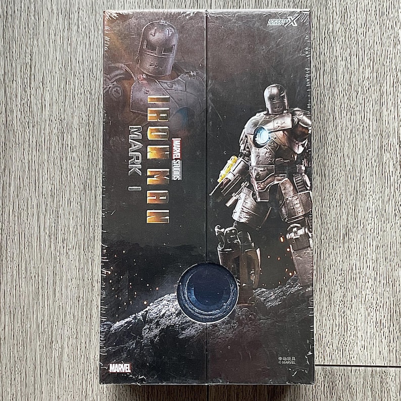 Może przedstawiać: Zapakowane pudełko z Iron Man Mark I z Marvel Studios. Opakowanie przedstawia wizerunki zbroi Iron Man w metalicznej kolorystyce. Logo Marvel jest widoczne w lewym dolnym rogu.