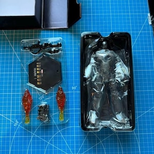 Może przedstawiać: Figurka Iron Mana w czarnym plastikowym pudełku ekspozycyjnym. Figurka jest srebrno-czarna, z akcesoriami, w tym bronią, podstawką z napisem "Iron Man Mark 1" i efektami płomieni.