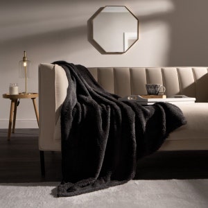 May include: A black faux fur throw blanket draped over a beige couch with a wooden tray and a cup on top. The couch is in a living room with a gold octagonal mirror and a lamp on a side table.