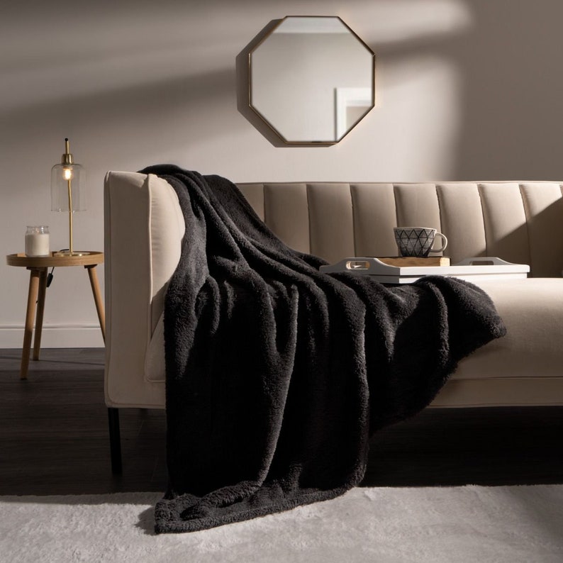 May include: A black faux fur throw blanket draped over a beige couch with a wooden tray and a cup on top. The couch is in a living room with a gold octagonal mirror and a lamp on a side table.