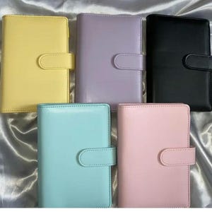 May include: Five colorful, leather-like binders in yellow, lavender, black, teal, and pink. Each binder has a rounded flap closure with a matching snap. The binders are arranged on a shiny, silver-colored surface.