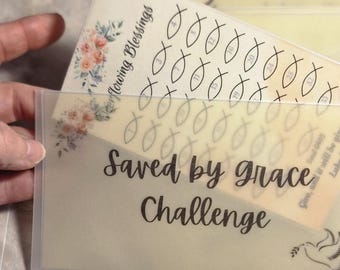 May include: A clear plastic envelope with the words "Saved by grace Challenge" in black script. Inside, a card with fish symbols and the words "Overflowing Blessings" and floral accents is visible. The envelope has three holes for binding.