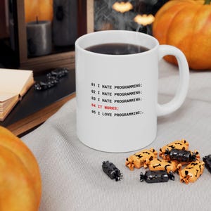 May include: A white ceramic mug featuring black text that reads 01 I HATE PROGRAMMING; 02 I HATE PROGRAMMING; 03 I HATE PROGRAMMING; 04 IT WORKS; 05 I LOVE PROGRAMMING; with the line 04 IT WORKS; printed in red ink on the front of the mug.