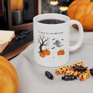 May include: A white ceramic mug featuring a graphic print with the text 'I'm here for the B00-leans;' accompanied by illustrations of a bare tree, two orange jack-o'-lanterns, a grey ghost, and several small black bats flying around.