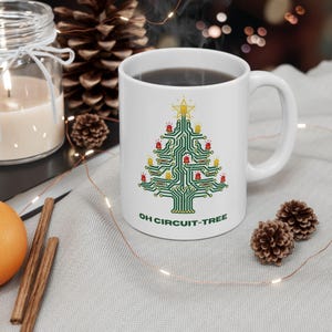 May include: A white ceramic mug featuring a graphic of a Christmas tree designed as a green circuit board with red and yellow LED lights and a star on top. The text "OH CIRCUIT-TREE" is printed in green capital letters below the tree design.
