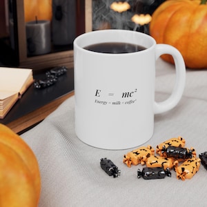 May include: A white ceramic mug featuring the black printed text E = mc2 and the subtext Energy = milk x coffee2. The mug has a standard handle and is filled with a dark liquid. It is positioned on a light-colored fabric surface with scattered candies.