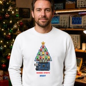 May include: A white long-sleeve sweatshirt featuring a graphic of a Christmas tree constructed from a breadboard with colorful LED lights and a gold star on top. Below the tree is a blue microcontroller board with the text MAKING SPIRITS BRIGHT.