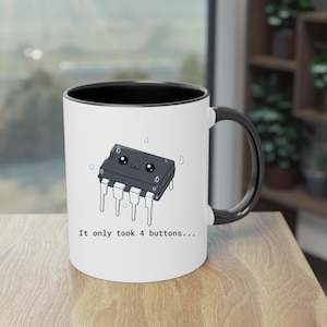 May include: A white ceramic mug with a black interior and handle, featuring a graphic of a black integrated circuit chip with a cute face and water droplets, accompanied by the text "It only took 4 buttons..." printed in a simple black font.