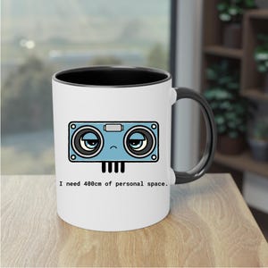 May include: A white ceramic mug with a black interior and handle. The front features a blue cartoon character with large, tired eyes and four small legs. Below the character, the text reads: I need 400cm of personal space.