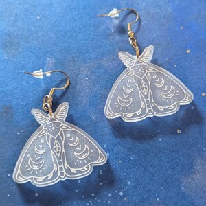 May include: A pair of clear acrylic moth-shaped earrings with intricate white line art. The earrings have gold-colored ear wires and are displayed against a dark blue background.