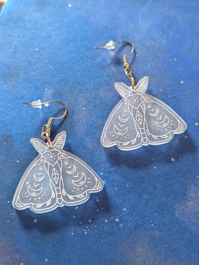 May include: A pair of clear acrylic moth-shaped earrings with intricate white line art. The earrings have gold-colored ear wires and are displayed against a dark blue background.