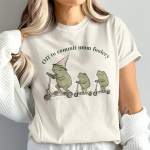 May include: A cream-colored short-sleeve t-shirt featuring a graphic of three green frogs riding scooters. The largest frog wears a pointed hat. Above the frogs, the text reads "Off to commit mom foolery" in a dark green, arched font.