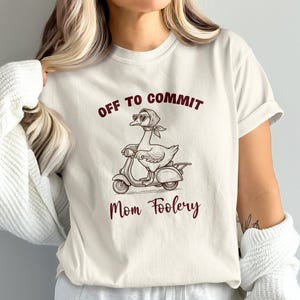 Puede incluir: Una camiseta de color crema con un gráfico de un ganso que lleva un pañuelo y gafas de sol mientras conduce un scooter. El texto sobre el ganso dice OFF TO COMMIT y el texto debajo dice Mom Foolery en una fuente de color rojo oscuro.