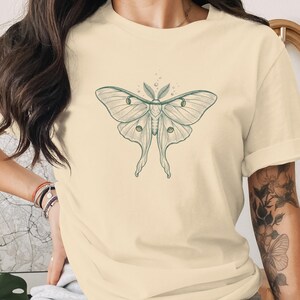 以下が含まれることがあります： クリーム色のTシャツ。羽を広げた蛾の精巧なイラストが描かれています。蛾は緑色で表現され、複雑な羽の模様と触角があります。カジュアルな普段着です。