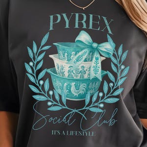 May include: Dark gray t-shirt featuring a teal graphic of Pyrex bowls, a bow, and the text "PYREX Social Club." The design is surrounded by a teal wreath and the phrase "IT'S A LIFESTYLE" is below the text.