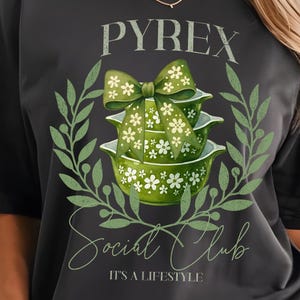 May include: Dark grey t-shirt with a green and white graphic of Pyrex bowls and a bow, encircled by a green wreath. The text "PYREX Social Club" and "IT'S A LIFESTYLE" are printed on the shirt.