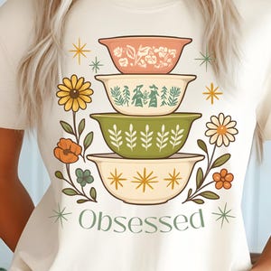 May include: Ivory-colored t-shirt featuring a stack of decorative bowls in shades of pink, green, and yellow, with floral accents and the word "Obsessed." The shirt is made of Comfort Colors 1717 material.
