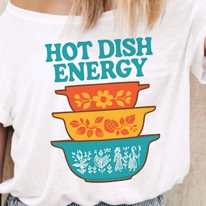 May include: White t-shirt with the text "HOT DISH ENERGY" in teal. Below the text are three stacked, colorful casserole dishes with floral and geometric designs. The image also includes the text "High Quality 300 dpi PNG Digital Download."