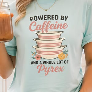 May include: A light blue t-shirt with the text "POWERED BY Caffeine AND A WHOLE LOT OF Pyrex" in pink and white. The shirt features a graphic of three stacked bowls. A glass jar with a straw is held in the hand.
