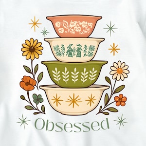 Puede incluir: Camiseta blanca con un diseño de cuatro cuencos decorativos en naranja, verde y amarillo. Los cuencos tienen diseños florales y geométricos. La palabra "Obsessed" está impresa debajo.