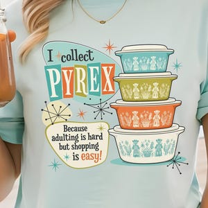 May include: A light blue t-shirt featuring a graphic of four stacked vintage-style bowls in teal, green, orange, and white. The text reads I collect PYREX Because adulting is hard but shopping is easy, surrounded by colorful starburst accents.