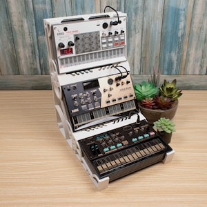 May include: A white rack holds several Korg Volca synthesizers and drum machines. The top unit displays "Korg Volca Sample" and a digital display. The rack is on a wooden surface with a blue and white wood-paneled backdrop. A small potted plant sits nearby.