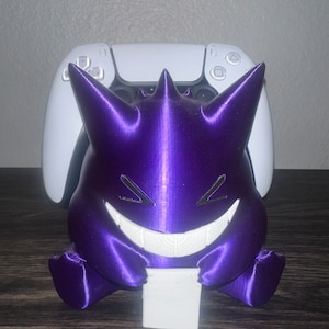 May include: A purple 3D-printed Gengar figure with a wide grin and spiky features, holding a white block. A white game controller is in the background. The figure is on a dark wooden surface.