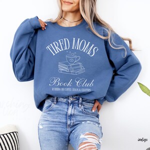 May include: Indigo blue sweatshirt with the text "TIRED MOMS Book Club" in white script. The design includes a coffee cup over a stack of books. The sweatshirt is worn with blue jeans.