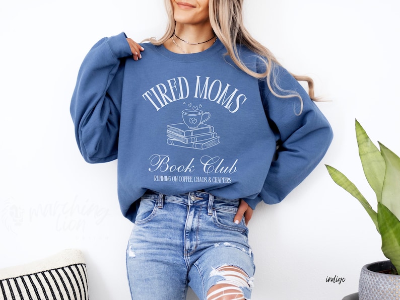 May include: Indigo blue sweatshirt with the text "TIRED MOMS Book Club" in white script. The design includes a coffee cup over a stack of books. The sweatshirt is worn with blue jeans.