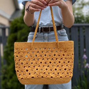 May include: A mustard yellow tote bag crafted from crocheted yarn, featuring a repeating floral motif pattern throughout the body, a solid rectangular base, and two thin, braided shoulder straps attached to the top edge of the bag.