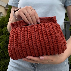May include: A rust-coloured, rectangular crochet clutch bag featuring a thick, ribbed texture and a built-in handle opening at the top. The bag is constructed from a dense, cord-like yarn with a consistent, repeating stitch pattern throughout the body.