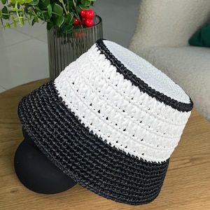 Könnte beinhalten: Ein gewebter Eimerhut in Schwarz und Weiß. Der Hut hat eine schwarze Krempe und ein weißes Band in der Mitte. Der Hut liegt auf einer schwarzen Kugel auf einer Holzoberfläche. Ein modisches Accessoire.