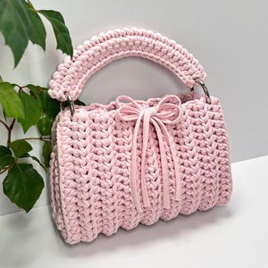 Könnte beinhalten: Hellrosa, handgehäkelte Handtasche mit rundem Griff und silbernen Metallringen. Die Tasche hat eine Schleife auf der Vorderseite und ein strukturiertes Stichmuster. Die Tasche hat eine weiche, pastellrosa Farbe.