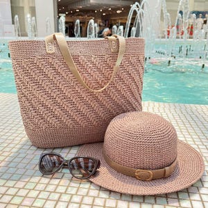 May include: A blush-coloured woven tote bag with tan leather handles sits next to a matching sun hat with a tan belt and buckle. A pair of dark sunglasses rests in front of the hat. The items are on a tiled surface.