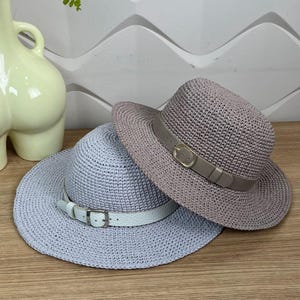 May include: Two woven sun hats. One is light blue with a white band and buckle, the other is mauve with a brown band and gold buckle. Both hats have a wide brim and are made of a textured material.