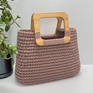 May include: A mauve-colored crochet tote bag with a light wood handle. The bag has a rectangular shape and a sturdy, woven texture. The handle is made of light-colored wood and is attached to the bag with matching mauve fabric.