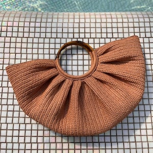 Könnte beinhalten: Eine gewebte, pfirsichfarbene Handtasche mit einem kreisförmigen Bambusgriff. Die Tasche hat ein plissiertes Design und eine abgerundete Form. Die Tasche liegt auf einer weiß-grauen Fliesenoberfläche.