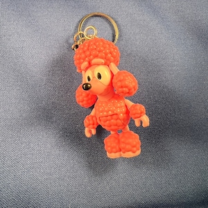 Puede incluir: Un llavero de plástico rosa en forma de caniche con un anillo de llave dorado. El caniche está hecho de pequeñas formas redondas y con forma de burbuja.