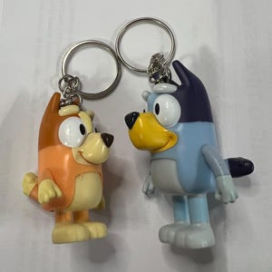 May include: Two plastic keychains featuring cartoon dogs from the animated series PAW Patrol. The keychain on the left is orange and brown with a yellow face and white eyes. The keychain on the right is blue with a yellow beak and white eyes.