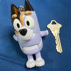 May include: A light purple Bluey figurine with black and brown ears, a black nose, and white accents. A silver key is next to the figurine. The figurine has a silver keychain attachment.