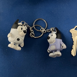 May include: Two cartoon dog keychains, one white and black, the other lavender and black, connected to a silver keyring. A silver key is next to the keychains. The keychains are on a blue surface.
