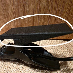 May include: Black foot pedal with a textured surface and a curved base. The pedal is connected to a black cable. The background is a wooden surface.