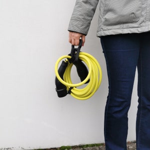 Könnte beinhalten: Ein gelbes Ladekabel für Elektrofahrzeuge mit einem schwarzen Griff und einem schwarzen Stecker an jedem Ende. Das Kabel ist aufgerollt und wird von einer Person gehalten.