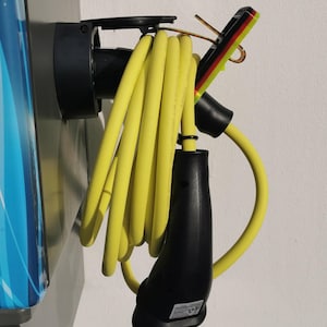 Puede incluir: Un cable de carga de vehículo eléctrico amarillo está enrollado y colgando de una estación de carga montada en la pared. El cable tiene un enchufe negro en el extremo y un gancho negro para sujetarlo en su lugar.